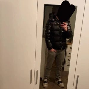Moncler Maya - Säljer en svart pufferjacka från Moncler med glansig finish och huva. Jackan har dragkedja framtill och två sidofickor med dragkedja. Perfekt för kalla dagar och riktigt snygg streetstil.