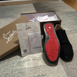 Louboutins - Säljer ett par svarta sneakers från Christian Louboutin med klassisk röd sula och svart snörning. Skorna har rund tå och är tillverkade i tyg med diskret logga bak på hälen. Kommer med originalkartong och kvitto.