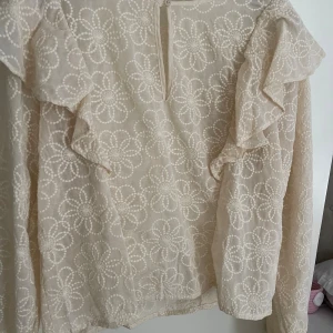 Beige blommig blus Carin Wester - Supersöt beige blus från Carin Wester med broderade blommor över hela tyget. Blusen har volangdetaljer på axlarna, lång ärm och knäppning i nacken. Perfekt för dig som gillar romantiska och luftiga plagg.