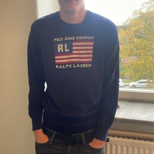 Blå tröja från Ralph Lauren - Säljer en mörkblå långärmad tröja från Ralph Lauren Polo Jeans Company med tryckt RL-logga och amerikansk flagga på bröstet. Tröjan har rund halsringning och ribbade muddar vid ärmslut och midja. Perfekt för dig som gillar klassisk streetstyle.