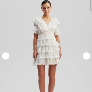 By malina klänning - Supersöt kort vit klänning med flera lager volanger och spetsdetaljer. Djup v-ringning och korta ärmar med volang ger en romantisk vibe. Perfekt för dig som vill sticka ut med en feminin och trendig look.