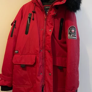 Röd parkajacka Simon and Son S - Röd parkajacka från Simon and Son med svart fuskpäls på huvan, flera dragkedjeförsedda fickor och snygga patchdetaljer på ärmen. Jackan har justerbar huva med snörning och stora fickor framtill. Perfekt för kalla dagar när du vill hålla stilen.