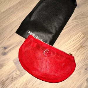 Röd kuvertväska från Giorgio Armani - Snygg röd kuvertväska från Giorgio Armani med halvmåneform och guldfärgad dragkedja. Väskan har diskret logga framtill och är tillverkad i glansigt syntetmaterial. Perfekt för att bära det viktigaste och ger en lyxig touch till din outfit.