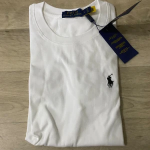 Vit t-shirt från Polo Ralph Lauren - Vit t-shirt från Polo Ralph Lauren med klassisk rund hals och korta ärmar. T-shirten är i mjuk bomull och har den ikoniska svarta Polo-loggan broderad på bröstet. Enkel och stilren design som funkar till allt.