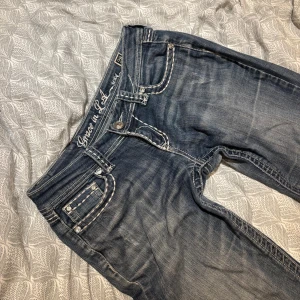 Lågmidjade bootcut jeans - Snygga blå jeans från Grace in LA med kontrastsömmar och broderade bakfickor med blingdetaljer. Låg midja och bootcut, klassisk femficksmodell. Jeansen har slitningar och dekorativa knappar på fickorna. Perfekta för dig som gillar detaljer och unik stil. De har två olika knappar på bilden och en liten fläck men det ser mer ut som en cool slitning (syns på bilderna). De är assnygga men tyvärr för stora för mig, skulle uppskatta jeansen till större S eller mindre M. Midjemått: 39, innerben: 80/81.