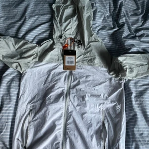 Vit vindjacka från The North Face - Snygg vit och ljusgrå vindjacka från The North Face med huva och hel dragkedja framtill. Jackan har två sidofickor med dragkedja och logga på bröstet. Perfekt för blåsiga dagar och outdoor-aktiviteter. Lätt och smidig att bära med sig.