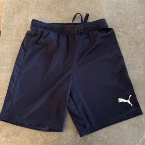 Marinblå träningsshorts från Puma - Säljer ett par marinblå träningsshorts från Puma med elastisk midja och vit logga på benet. Lätta och sköna med sportig look, perfekta för gymmet eller löprundan. Tillverkade i syntetmaterial som andas.