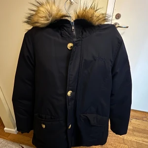 Svart parkasjacka från Svea med fuskpäls - Säljer en svart parkasjacka från Svea med snygg fuskpäls på huvan, guldiga knappar och broderad amerikansk flagga på ärmen. Jackan har dragkedja och stora fickor framtill, perfekt för kalla dagar.