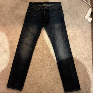 Lee W33 L34 mörkblå - Mörkblå Lee jeans med snyggt slitna detaljer och klassisk femficksdesign. Jeansen har raka ben och kontrastsömmar, vilket ger en stilren look. Perfekta för dig som gillar en tidlös och clean stil.