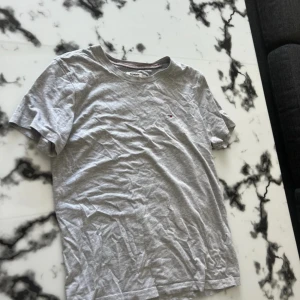 Grå t-shirt från Tommy Hilfiger - En stilren grå t-shirt från Tommy Hilfiger med liten broderad logga på bröstet. Klassisk rund halsringning och korta ärmar. Tillverkad i mjuk bomull som känns skön mot huden. Perfekt för en avslappnad look.