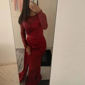 Röd långklänning med paljetter - Säljer en elegant röd långklänning med figurnära passform och långa ärmar. Överdelen är täckt av glittriga paljetter och har en rund halsringning. Kjolen är slät med drapering och ett långt släp som ger extra drama. Perfekt för bal eller fest!