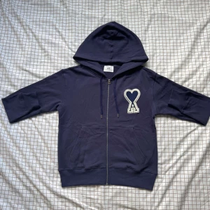 Mörkblå hoodie från Ami Paris med dragkedja - Säljer en mörkblå hoodie från Ami Paris med dragkedja och huva. Tröjan har ett stort broderat hjärta med bokstaven A på bröstet, ribbade muddar och fickor framtill. Tillverkad i mjuk bomull och perfekt för en chill look.