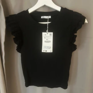 Svart volangtopp  - Svart topp från Zara med volangdetaljer på axlarna. Stilren och söt. Helt oanvänd med prislappen kvar! 🤗