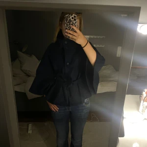 Mörkblå kappa med vida ärmar - Säljer en mörkblå kappa med hög krage och tre stora knappar framtill. Jackan är i nyskick!