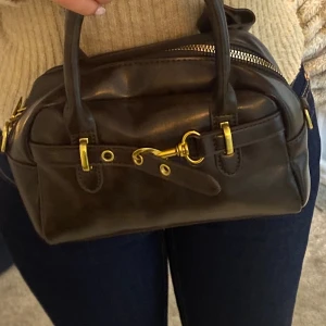 Brun handväska i skinn med gulddetaljer - Snygg brun handväska i skinn med guldfärgade metalldetaljer och dragkedja. Väskan har två handtag och avtagbar axelrem, samt dekorativt spänne framtill. Kompakt och stilren modell som ger en lyxig känsla.