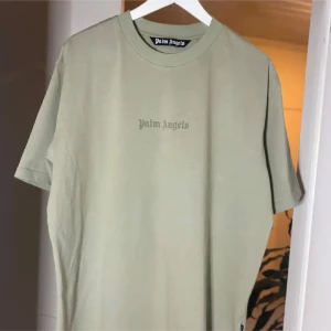 Palm Angels beige t-shirt - Snygg beige t-shirt från Palm Angels med logga tryckt på bröstet och stort tryck på ryggen. Klassisk rund hals och avslappnad passform. Perfekt för dig som gillar streetwear och vill ha en clean look med en touch av lyxmärke.