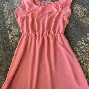 Söt rosa volangklänning - En superfin rosa kortklänning med volangdetaljer vid halsringningen och ärmlös design. Klänningen har markerad midja och ett lätt, flowy tyg som ger en luftig känsla. Perfekt för varma dagar och somriga tillfällen.