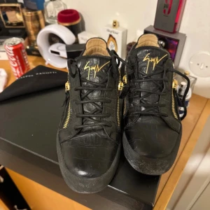 Svarta sneakers från Giuseppe Zanotti - Säljer ett par svarta sneakers från Giuseppe Zanotti i lyxigt skinn med krokodilpräglad yta. Skorna har guldfärgade dragkedjor på sidorna, svart snörning och den ikoniska loggan i guld på plösen. Riktigt snygga och unika skor för dig som vill sticka ut.