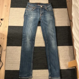 Nudie Jeans Grim Tim blå jeans 28/32 - Snygga Nudie Jeans Grim Tim i klassisk blå tvätt med orangea sömmar och slitningar på fickorna. Modellen har rak passform och normal midja. Tillverkade av 98% ekologisk bomull och 2% elastan för skön stretch. Perfekt för dig som gillar stilrena jeans med lite edge.