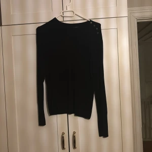 Svart långärmad tröja från ONLY - Svart långärmad tröja från ONLY med rund hals och snygga knappar på ena axeln. Tröjan är i en smal passform och tillverkad i mjukt material som känns skönt mot huden. Perfekt att styla med jeans eller kjol för en clean look.