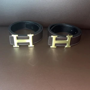  Brunt bälte med guldspänne - Säljer ett stilrent brunt bälte med ikoniskt H-spänne i guld. Bältet är tillverkat i exklusivt skinn och har vit kontrastsöm längs kanterna. Perfekt accessoar för att lyfta din outfit med en lyxig touch.