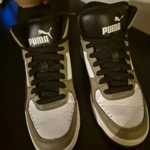 Puma high-top sneakers svart/vit - Snygga high-top sneakers från Puma i svart, vitt och olivgrönt. Skorna har klassisk snörning, rund tå och platt sula. Puma-logga syns både på plösen och hälen. Tillverkade i syntetmaterial med skinnliknande yta. Perfekta för dig som gillar sportig streetstil.