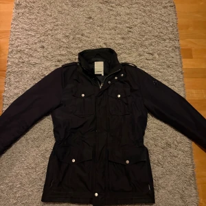 Svart parkajacka från J.Lindeberg - Svart parkajacka från J.Lindeberg med huva, dragkedja och tryckknappar. Jackan har fyra stora fickor framtill och justerbar midja. Perfekt för kyliga dagar och har en stilren look med diskreta detaljer.