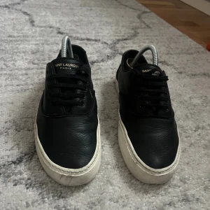 Svarta sneakers från Saint Laurent - Svarta sneakers från Saint Laurent Paris i skinn med vit platt sula och klassisk snörning. Låg modell med rund tå och diskret logga på plösen. Perfekta för en clean och stilren look.