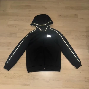 Svart EA7 hoodiejacka med reflexdetaljer - Svart hoodiejacka från EA7 Emporio Armani med dragkedja och reflekterande detaljer längs ärmar och huva. Jackan har en stilren look med EA7-logga på bröstet och är tillverkad i mjukt syntetmaterial. Perfekt för dig som gillar sportig och clean stil.