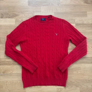 Röd kabelstickad tröja från Gant - Säljer en röd kabelstickad tröja från Gant med rund halsringning och diskret logga på bröstet. Tröjan har långa ärmar och ribbade muddar vid ärmslut och nederkant. Perfekt för dig som gillar klassisk och stilren look.