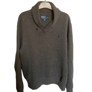 Ralph lauren cardigan - Snygg grå stickad tröja från Polo Ralph Lauren med klassisk krage och knappar vid halsen. Tröjan har långärmad passform och den ikoniska broderade loggan i mörkblått på bröstet. Perfekt för dig som gillar stilrena och tidlösa plagg.