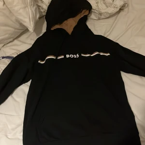 Svart hoodie från BOSS med beige detaljer - Svart hoodie från BOSS med beige insida på huvan och coola ränder i vitt och beige över bröstet. Stort BOSS-tryck framtill. Perfekt för dig som gillar stilren streetwear med premiumkänsla.