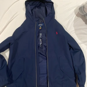 Blå vindjacka från Polo Ralph Lauren - Snygg marinblå vindjacka från Polo Ralph Lauren med huva och dragkedja. Jackan har två stora fickor framtill och ikonisk röd logga på bröstet. Tillverkad i polyester, perfekt för blåsiga dagar. Enkel och stilren design med elastiska muddar vid ärmslut.