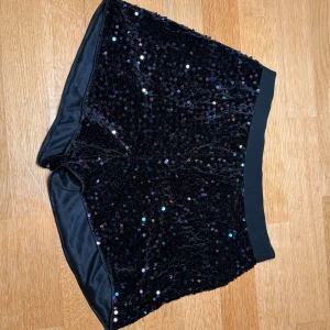 Svarta paljettshorts med elastisk midja - Glittriga svarta shorts med massor av paljetter som reflekterar i regnbågens färger. De har en mjuk, elastisk midja och är tillverkade i ett sammetsliknande material som känns lyxigt. Perfekta för dig som vill sticka ut och älskar att festa:)