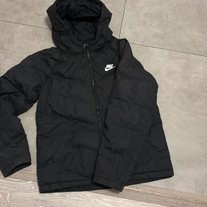 Svart pufferjacka från Nike XL - Säljer en svart pufferjacka från Nike i storlek XL. Jackan har huva, dragkedja framtill och klassisk Nike-logga på bröstet. Perfekt för kyliga dagar och enkel att matcha med allt. Snygg och sportig look som alltid funkar.158/170