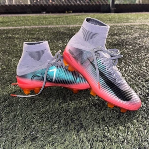 Nike Mercurial CR7 fotbollsskor - Säljer ett par Nike Mercurial CR7 fotbollsskor med högt skaft och snörning. Skorna har en snygg färgskiftning i silver, svart och röd med orange dobbar. Tillverkade i syntetmaterial för bästa grepp och känsla på planen. Perfekta för dig som vill sticka ut på fotbollsplanen. Pris kan diskuteras.