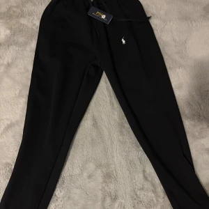 Svarta mjukisbyxor Polo Ralph Lauren - Svarta mjukisbyxor från Polo Ralph Lauren med klassisk vit logga broderad på benet. Byxorna har elastisk midja med snörning och är gjorda i mjukt bomullsmaterial. Perfekta för chill eller streetwear och har en clean, stilren look.