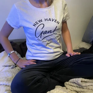 Vit Gant t-shirt med tryck - Vit t-shirt från Gant med svart tryck på bröstet där det står 'New Haven Gant EST. 1949 NHCT U.S.A.'. Klassisk rund halsringning och korta ärmar. Skön och mjuk i bomull, perfekt till jeans eller shorts.