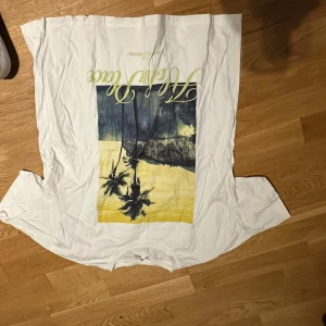 Vit t-shirt med palmmotiv från Pull&Bear - Vit t-shirt från Pull&Bear i bomull med tryck av palmer och strand i blått och gult framtill. Klassisk passform med rund hals och korta ärmar. Perfekt för en chill sommarlook.