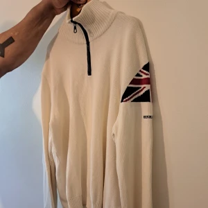 Vit stickad tröja Henri Lloyd XXL - Vit stickad tröja från Henri Lloyd med half zip och hög krage. Union Jack-detalj i rött, blått och vitt på ena ärmen samt broderad logga. Ribbad vid krage, ärmslut och nederkant. Storlek XXL. 