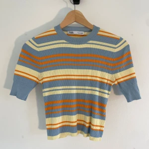 Randig ribbad topp från Zara - Säljer en ribbad topp från Zara med korta ärmar och rund hals. Toppen har ett färgglatt randigt mönster i ljusblått, gult och orange. Materialet är mjukt och stretchigt, perfekt för dig som gillar retrovibbar och vill sticka ut med coola färger.