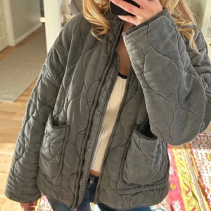 Grå quiltad jacka från Zara - Säljer en grå quiltad jacka från Zara med oversized passform. Jackan har två stora fickor framtill, dragkedja och en hög krage. Materialet är manchester och jackan har ett vågigt quiltat mönster som ger en cool look. Perfekt för höst och vinter.