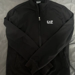 EA7 tröja svart  - Svart j från EA7 Emporio Armani med vit logga på bröstet och diskreta vita detaljer på ryggen. Tröjan har hel dragkedja framtill, ståkrage och långa ärmar. Snygg och stilren design som passar perfekt till streetwear. Den är storlek M men sitter mer som S