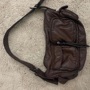 Brun axelväska i skinn från urban outfitters - Säljer en brun axelväska från urban outfitters i mjukt skinn med flera praktiska fickor och justerbar rem. Väskan har coola detaljer med dragkedjor och en snygg, lite slouchy form som passar perfekt till en avslappnad stil.