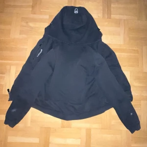 Svart fleecejacka från Sail Racing - Svart fleecejacka från Sail Racing med huva och dragkedja framtill. Jackan har broderad logga på bröstet och praktiska fickor med dragkedja. Perfekt för kyliga dagar och enkel att matcha med allt.