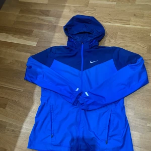Blå Nike vindjacka med huva - Nike vindjacka i två nyanser av blått med huva och dragkedja framtill. Jackan har två fickor med dragkedja och en vit Nike-logga på bröstet. Perfekt för blåsiga dagar och sportiga outfits.