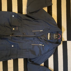 Mörkblå fieldjacka, Morris - Snygg mörkblå fieldjacka från Morris med klassisk krage, stora fickor med knappar och dragkedja framtill. Jackan har en stilren look med diskreta detaljer och är tillverkad i polyester. Perfekt för dig som gillar en clean och tidlös stil.