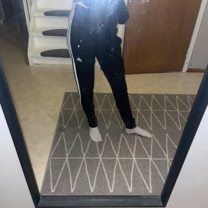 Svarta Adidas mjukisbyxor med vita ränder - Svarta mjukisbyxor från Adidas med klassiska tre vita ränder längs sidorna och Adidas-logga på låret. Byxorna har hög midja, snörning i midjan och är tillverkade i mjuk bomullsmix. Perfekta för chill eller träning.