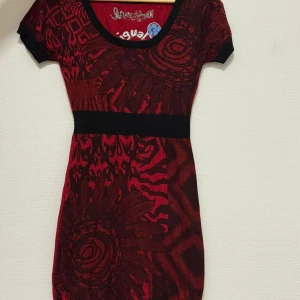 Röd mönstrad kortklänning Desigual - Unik röd och svart kortklänning från Desigual med coola grafiska mönster och stora blommor framtill. Klänningen har rund hals, korta ärmar och markerad midja med svart band. Perfekt för dig som gillar färg och mönster!