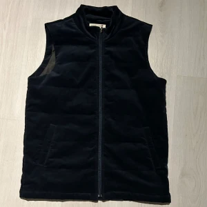 Mörkblå manchesterväst - Snygg mörkblå väst från sayless i manchester med rak passform och hög krage. Västen har två sidofickor och stängs med dragkedja framtill. Insidan är fodrad i slätt material för extra komfort. Perfekt att slänga över en hoodie när det är kyligt ute. Hör av er med prisförslag eller om ni vill ha fler bilder eller mått